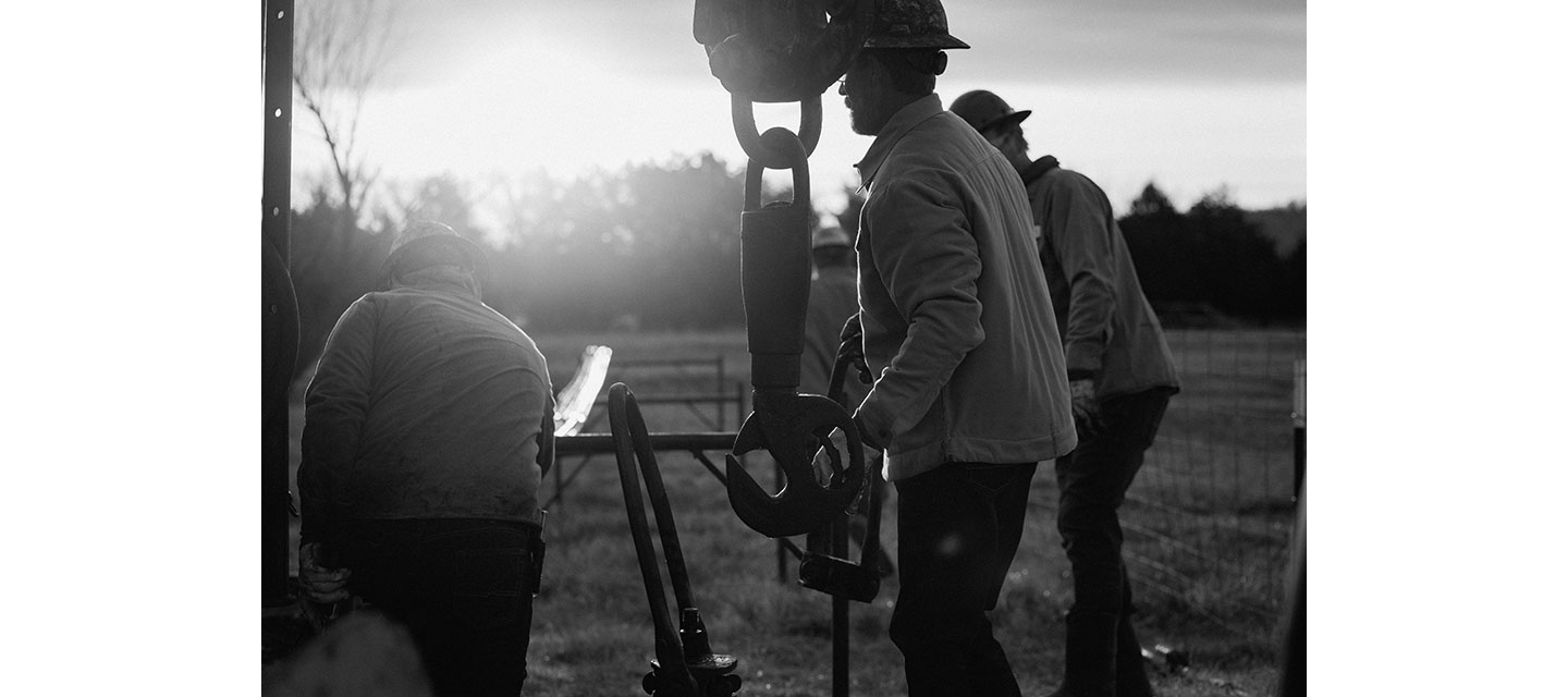 Varios trabajadores con cascos y chaquetas operan maquinaria pesada al aire libre al amanecer o al atardecer. Un trabajador maneja un gancho industrial grande, mientras otros ayudan en una zona cercada con césped.